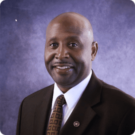 Professional portrait of a smiling man in a suit against a purple backdrop.